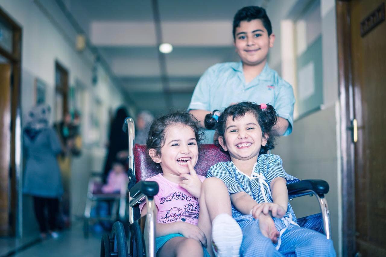 Two smiling girls sharing a wheelchair at Narayana Health City